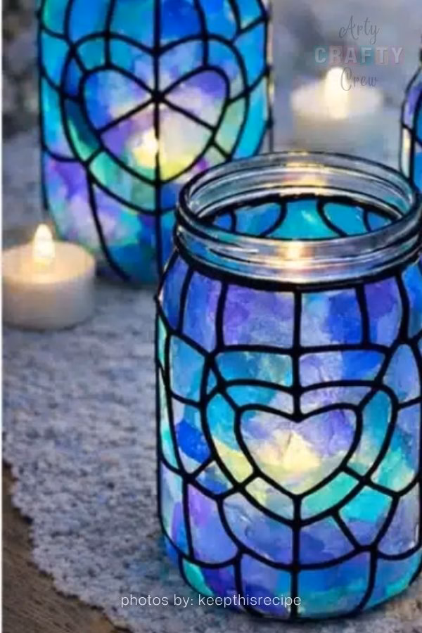 A creative display of three glass jars upcycled with colorful crochet covers. The jars are decorated with various patterns, including vibrant stripes and a floral motif, and are being used as functional holders for crochet hooks and craft supplies. This image is part of a crochet roundup from https://artycraftycrew.com/.