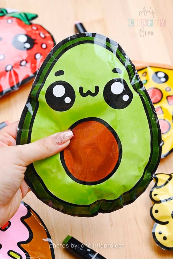 A hand holds a large, handmade paper squishy shaped like a green avocado with a brown pit and a cute, smiling kawaii face. The squishy is laminated with clear tape for a glossy finish, part of a DIY roundup from https://artycraftycrew.com/.