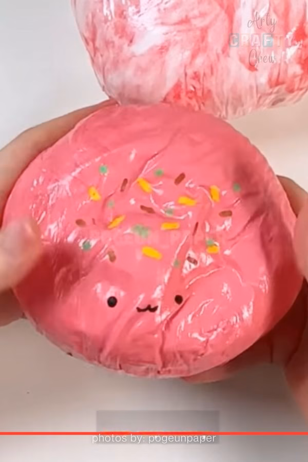 A close-up of a handmade paper squishy shaped like a round donut with bright pink frosting, colorful sprinkles, and a small, cute face. The craft is being held and squeezed by two hands, featured on https://artycraftycrew.com/.