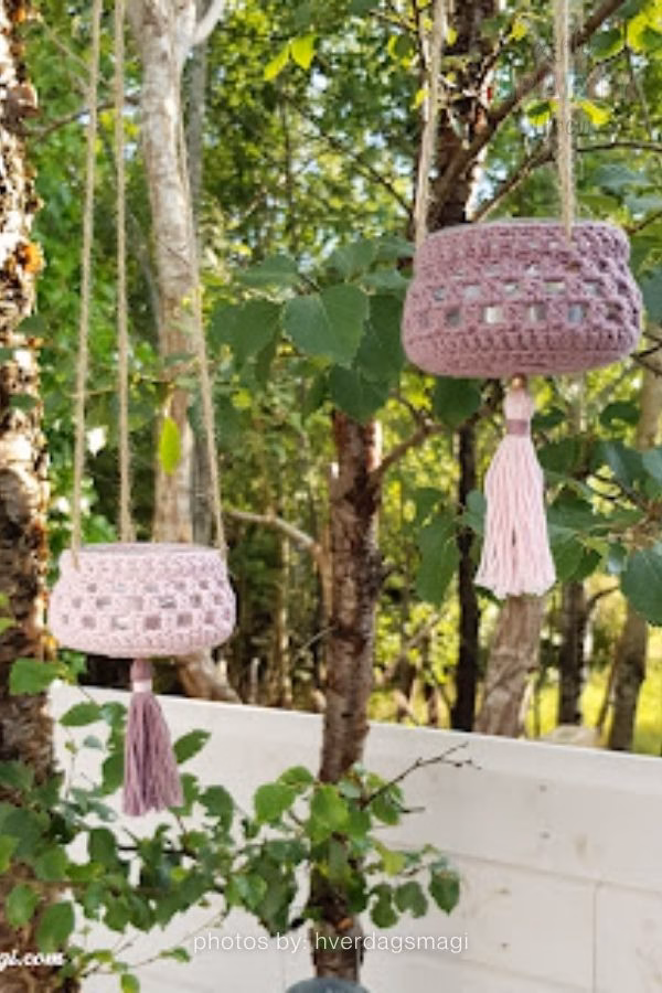 A close-up of a glass jar lantern featuring a colorful, textured crochet cover with stripes of vibrant pink, orange, green, and blue. The lantern is hanging outdoors against a soft, out-of-focus background of green trees. This image is part of a crochet roundup from https://artycraftycrew.com/.