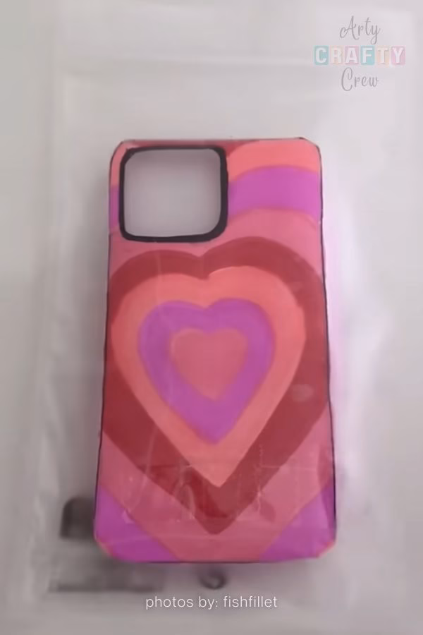 A handmade paper craft designed to look like a phone case, featuring a series of concentric hearts in shades of pink and purple. This DIY desk decor idea is part of a roundup from https://artycraftycrew.com/.