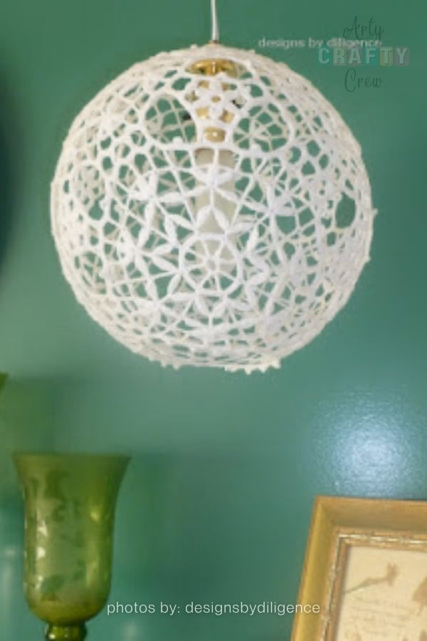 A glass jar lantern with a beautifully textured crochet cover in shades of deep teal and blue. The jar is hanging outdoors by a wire handle, creating a simple and charming DIY lighting piece for a garden. This image is part of a crochet roundup from https://artycraftycrew.com/.