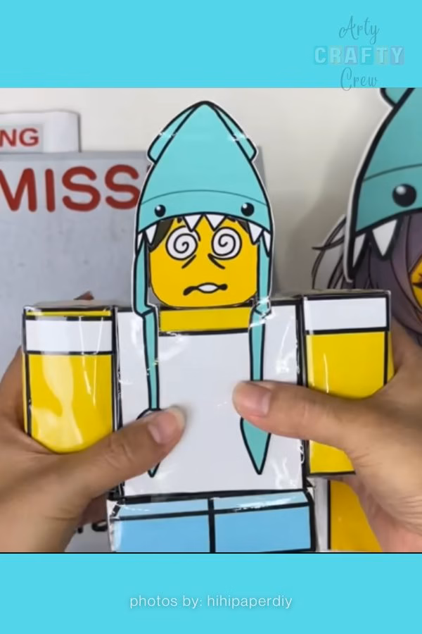 A hand holds a rectangular paper craft character inspired by Roblox, wearing a light blue shark hat and featuring a dizzy expression with swirl eyes. This beginner-friendly paper project is from https://artycraftycrew.com/.