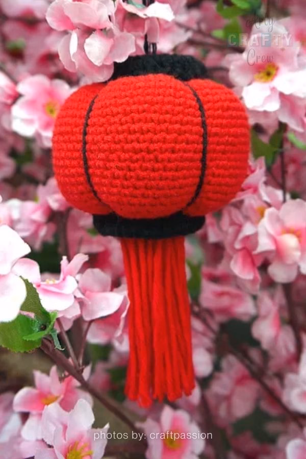 A close-up of a glass jar repurposed as a decorative lantern, featuring a vibrant pink and orange crochet sleeve with an open-stitch texture. The lantern is hanging outdoors against a soft, natural background of green leaves and garden foliage. This image is part of a crochet roundup from https://artycraftycrew.com/.
