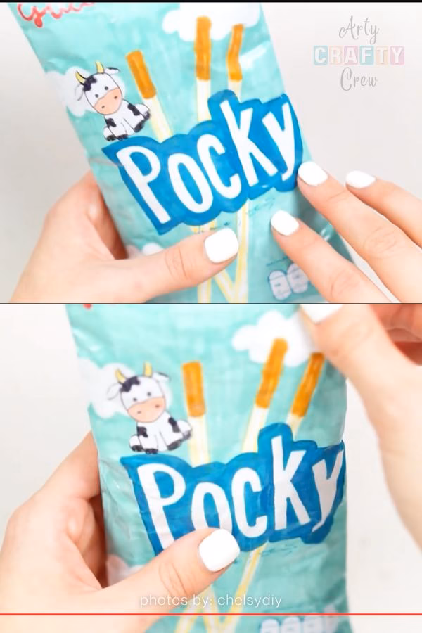 Two hands with white nail polish squeeze a light blue handmade paper squishy designed to look like a bag of Pocky sticks, featuring a cute cow illustration and biscuit stick graphics. Shared as part of a DIY collection on https://artycraftycrew.com/.