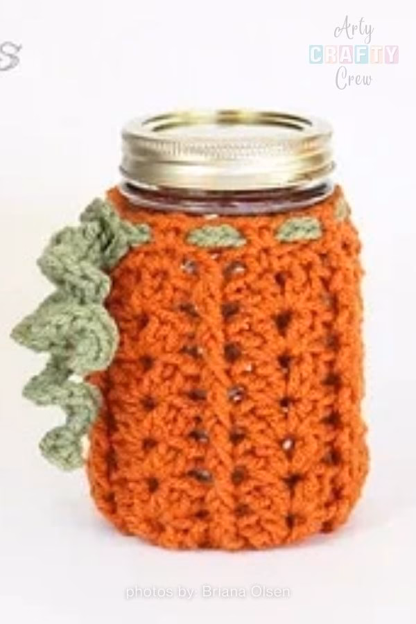 A close-up of a glass jar lantern with a white, intricate crochet cover. The jar is hanging by a wire handle against a blurred green background, showcasing a simple and elegant DIY lighting idea for a garden. This image is part of a crochet roundup from https://artycraftycrew.com/.