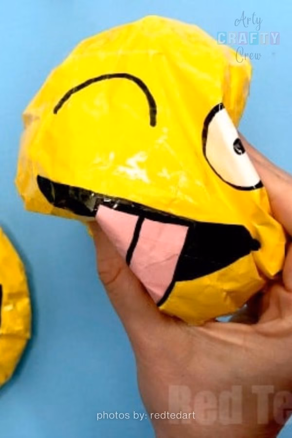 A close-up of a bright yellow handmade paper squishy featuring a winking emoji face with its tongue sticking out. The texture is crinkled and shiny from clear tape lamination, featured in a craft collection from https://artycraftycrew.com/.