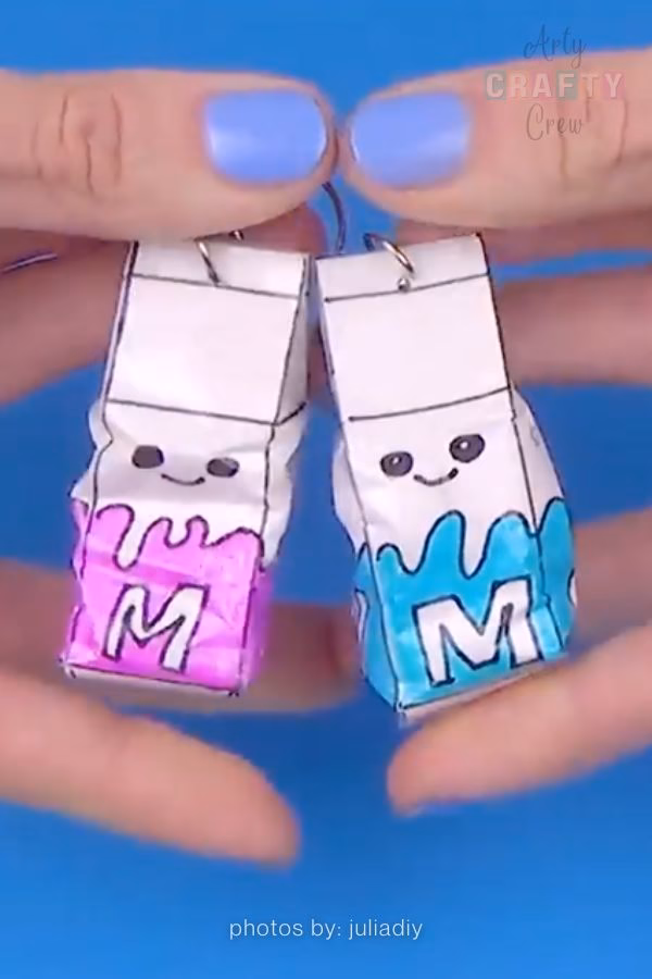 Two miniature handmade paper squishies shaped like milk cartons, one with pink accents and one with blue. Both feature tiny smiling faces and are shown being held by fingers with blue nail polish, featured on https://artycraftycrew.com/.