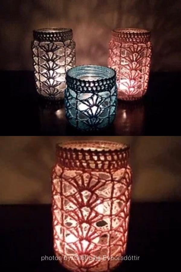 A close-up of two glass jar lanterns hanging outdoors against a background of green garden foliage. The jars are covered in textured crochet sleeves, one featuring warm sunset tones like orange and pink, and the other in cool shades of teal and blue. This image is part of a crochet roundup from https://artycraftycrew.com/.