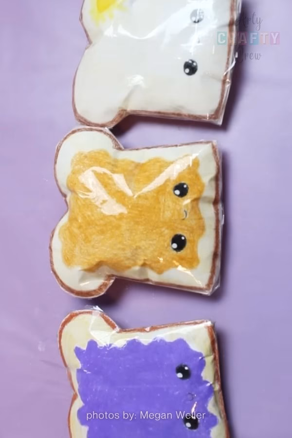 A vertical arrangement of three handmade paper squishies shaped like slices of bread. One slice is plain, one has yellow spread, and one has purple jam, all featuring cute kawaii eyes. These are part of a craft collection from https://artycraftycrew.com/.