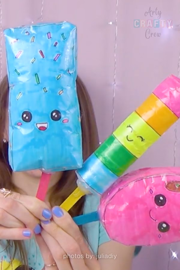 A person holds three handmade popsicle-shaped paper squishies. One is a blue rectangular popsicle with sprinkles, one is a multi-colored striped popsicle, and one is a pink oval popsicle, all featuring cute faces. Part of a low-budget DIY roundup from https://artycraftycrew.com/.