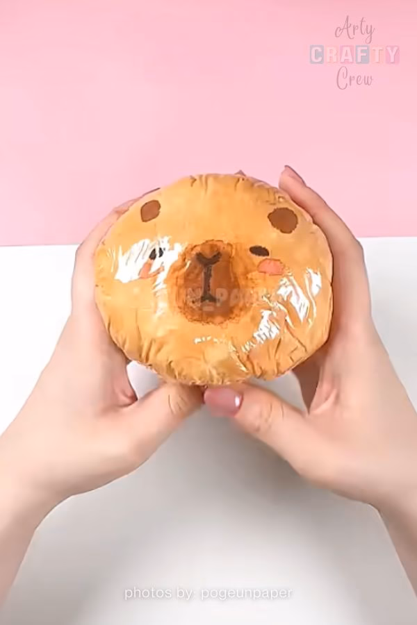 A top-down view of a round, handmade paper squishy designed to look like a capybara's face. Two hands are holding the tan-colored, glossy-textured craft. This creative project is shared by https://artycraftycrew.com/.