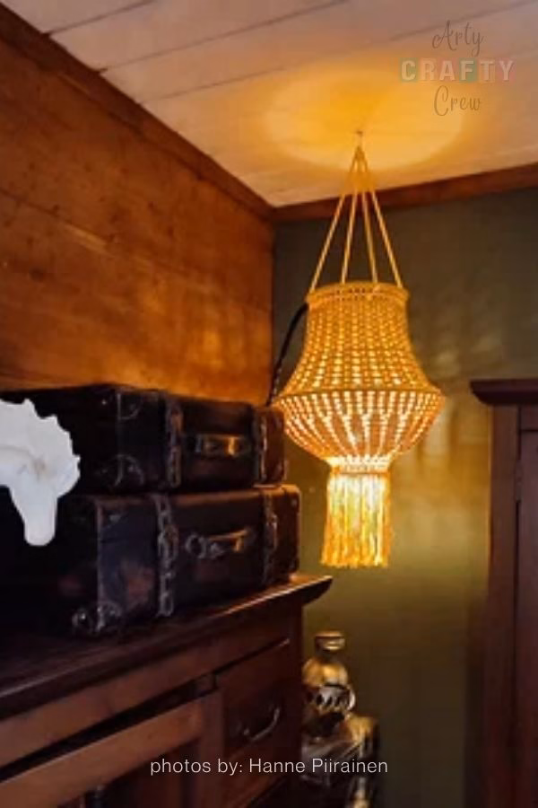 A bright glass jar lantern with a textured, teal-colored crochet cover hanging outdoors. The jar features a sturdy wire handle and is positioned against a background of lush green trees, perfect for a summer evening. This image is part of a crochet roundup from https://artycraftycrew.com/.