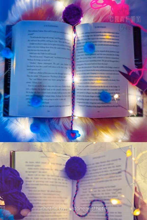 A split image featuring a handmade pompom bookmark. The top half shows an open book illuminated by fairy lights with blue pompoms scattered around. The bookmark, a dark purple pompom on a braided, multi-colored string, rests inside. The bottom half shows a person holding the book and the dark purple pompom bookmark, with a skein of dark purple yarn visible. Fairy lights are also present. The text "photos by: justoneaesthete" is at the bottom. This is part of a craft roundup from https://artycraftycrew.com/.