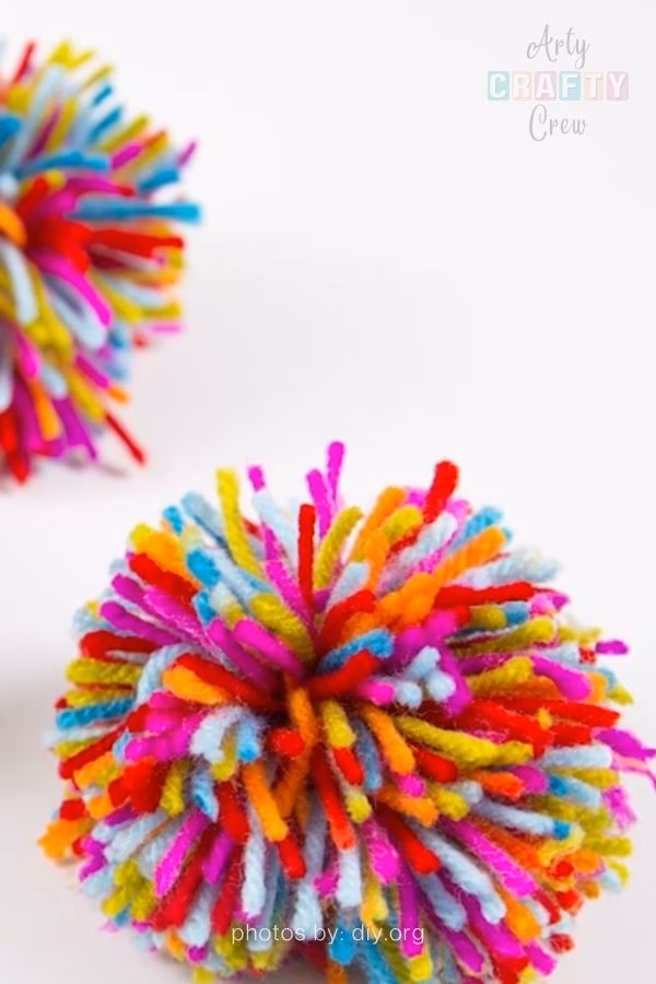 A close-up photograph of a vibrant, multi-colored yarn pompom resting on a white surface. The pompom is made of distinct, shaggy strands of bright pink, red, orange, yellow, and blue yarn. A second similar pompom is partially visible and out of focus in the background to the left. The Arty Crafty Crew logo is visible in the top right corner, and text at the bottom reads "photos by: diy.org". This is part of a crochet roundup from https://artycraftycrew.com/.
