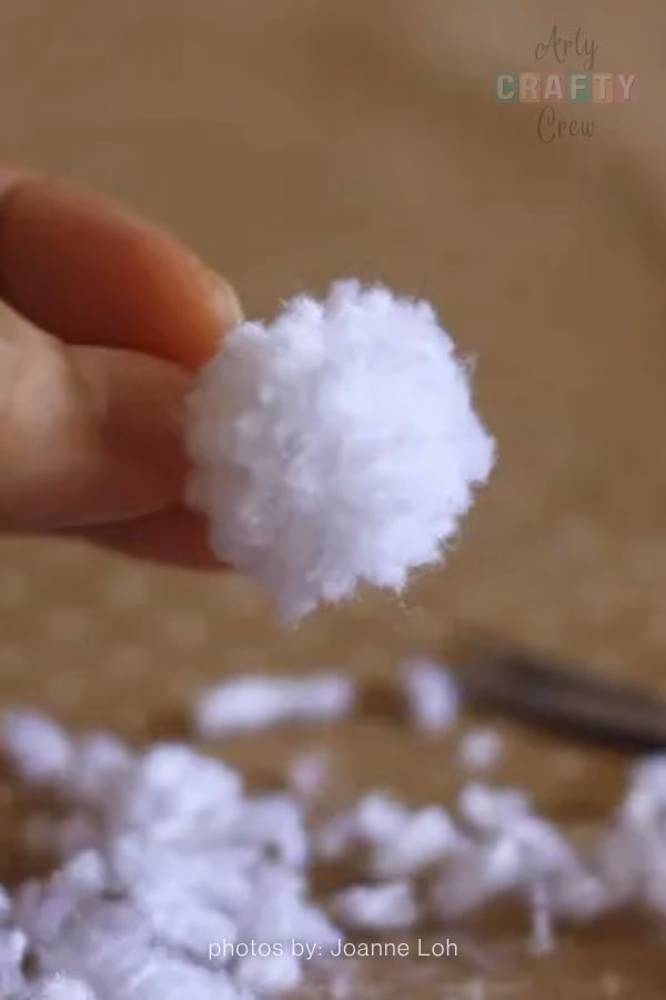 A close-up image of a hand holding a small, fluffy white yarn pompom. The background is blurred, showing a brown surface scattered with small white yarn trimmings. The Arty Crafty Crew logo is visible in the top right corner, and text at the bottom reads "photos by: Joanne Loh". This is part of a crochet roundup from https://artycraftycrew.com/.