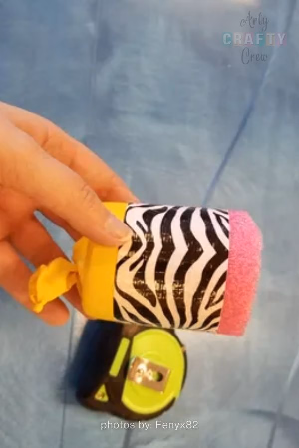A close-up photograph of a hand holding a DIY pompom shooter against a blue surface. The shooter is made from a short tube wrapped in black and white zebra print tape, featuring a stretched and tied yellow balloon on one end and a pink foam ring on the open end. A yellow and black tape measure is partially visible in the blurred background below. The Arty Crafty Crew logo is visible in the top right corner, and text at the bottom reads "photos by: Fenyx82". This is part of a craft roundup from https://artycraftycrew.com/.