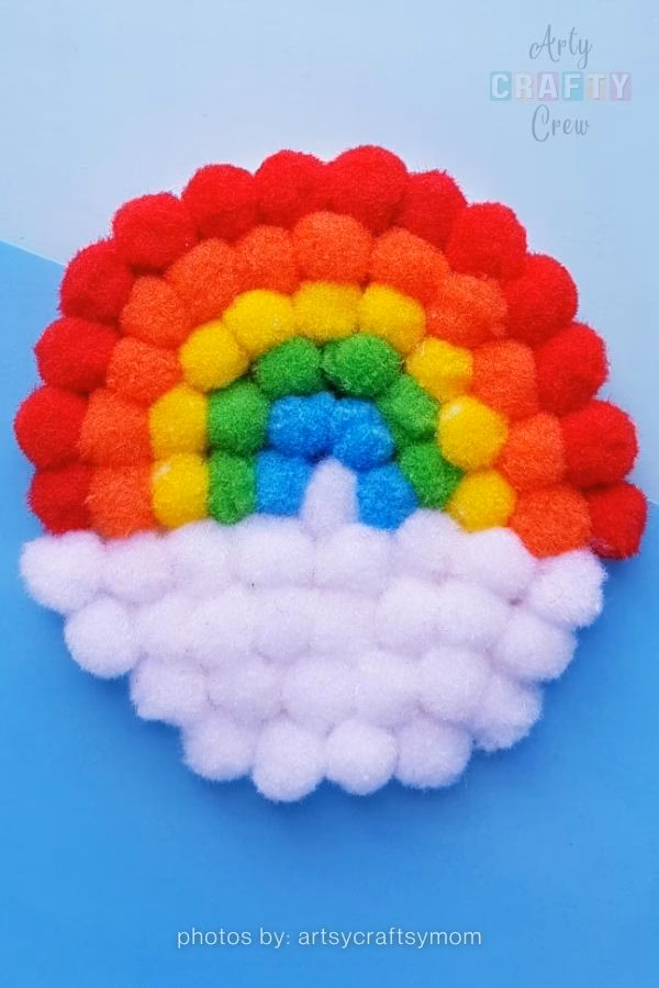 A craft project featuring a rainbow made of small, tightly packed pompoms in red, orange, yellow, green, and blue, with a fluffy white pompom cloud at the base. It is placed against a two-toned blue background. Text at the bottom reads "photos by: artsycraftsymom" and the Arty Crafty Crew logo is in the top right corner. This is part of a craft roundup from https://artycraftycrew.com/.