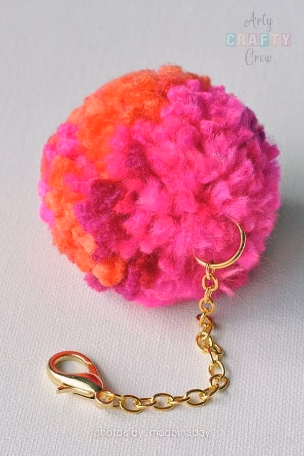 A close-up photograph of a handmade pompom keychain resting on a white surface. The large, fluffy pompom features patches of bright pink and vibrant orange yarn. It is attached to a short gold-colored metal chain with a lobster clasp at the end. The Arty Crafty Crew logo is visible in the top right corner, and text at the bottom reads "photos by: madeinaday". This is part of a craft roundup from https://artycraftycrew.com/.