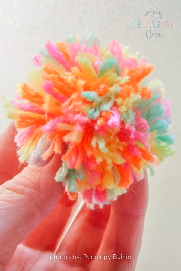 A close-up image showing a hand holding a brightly colored, multi-toned yarn pompom against a plain, light background. The pompom is made of shaggy yarn pieces in pastel green, bright orange, bright pink, and light yellow. The Arty Crafty Crew logo is visible in the top right corner, and text at the bottom reads "photos by: Penolopy Bulnic". This is part of a crochet roundup from https://artycraftycrew.com/.