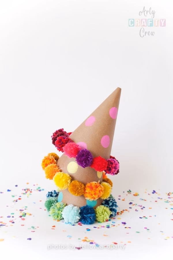 A photograph of a brown paper party hat resting on a white surface scattered with small, colorful confetti. The hat is decorated with round stickers and three distinct rings of yarn pompoms: the top ring features pink and purple pompoms with pink stickers; the middle ring has yellow and orange pompoms with yellow stickers; and the bottom ring consists of blue and green pompoms with teal stickers. The Arty Crafty Crew logo is visible in the top right corner, and text at the bottom reads "photos by: tellloveandparty". This is part of a crochet roundup from https://artycraftycrew.com/.
