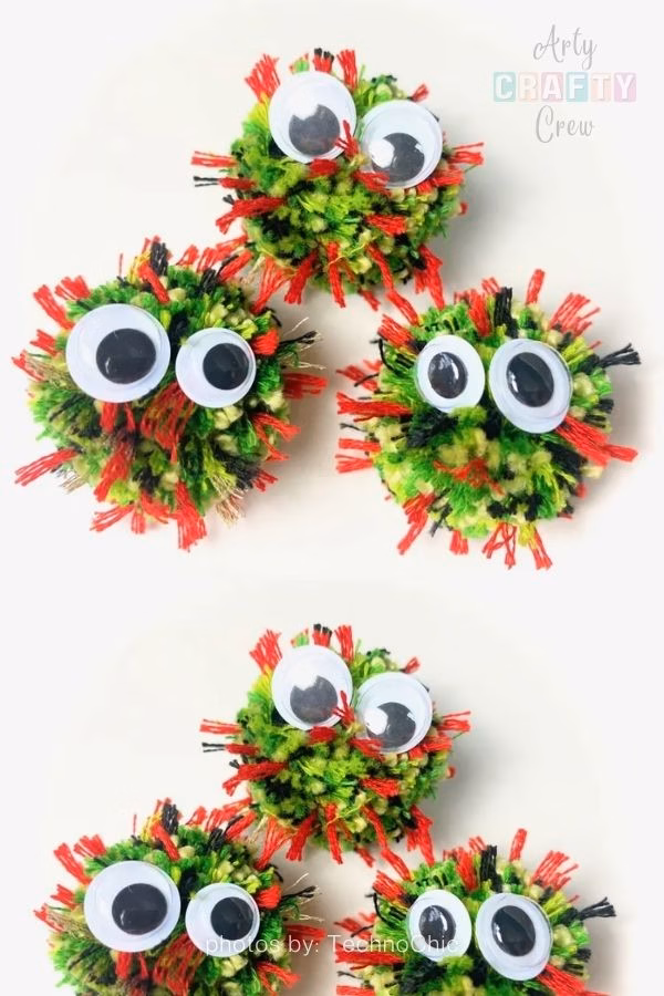 A split image showing three small, handmade pompom monsters resting on a plain white surface. The pompoms are crafted from green, yellow, and black yarn, with spiky red yarn pieces sticking out irregularly. Each monster has two large, mismatched googly eyes attached to it. The Arty Crafty Crew logo is visible in the top right corner, and text at the bottom reads "photos by: TechnoChic". This is part of a craft roundup from https://artycraftycrew.com/.