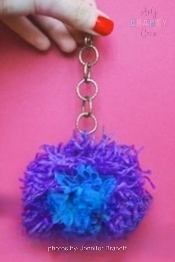 A hand with bright red nail polish holds up a decorative keychain against a solid bright pink background. The keychain features a fluffy, two-toned yarn pompom&mdash;predominantly purple with a prominent patch of bright blue in the center&mdash;attached to a bronze-colored metal chain with circular links. The Arty Crafty Crew logo is visible in the top right corner, and text at the bottom reads "photos by: Jennifer Branett". This is part of a craft roundup from https://artycraftycrew.com/.
