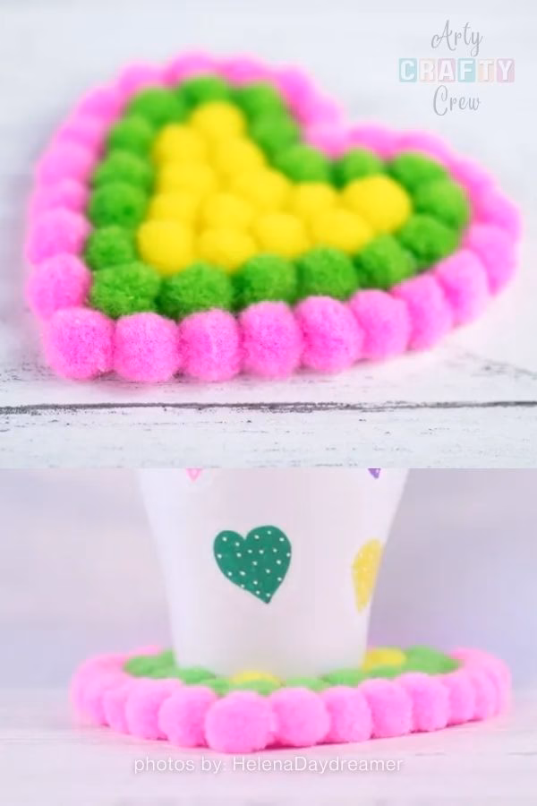 A split image demonstrating a handmade pompom coaster. The top half features a close-up of the heart-shaped coaster, created with a bright yellow pompom center, a vibrant green middle layer, and a neon pink outer edge, resting on a distressed white wooden surface. The bottom half shows the coaster in use, holding a white cup decorated with matching green and yellow heart decals. The Arty Crafty Crew logo is visible in the top right corner, and text at the bottom reads "photos by: HelenaDaydreamer". This is part of a craft roundup from https://artycraftycrew.com/.