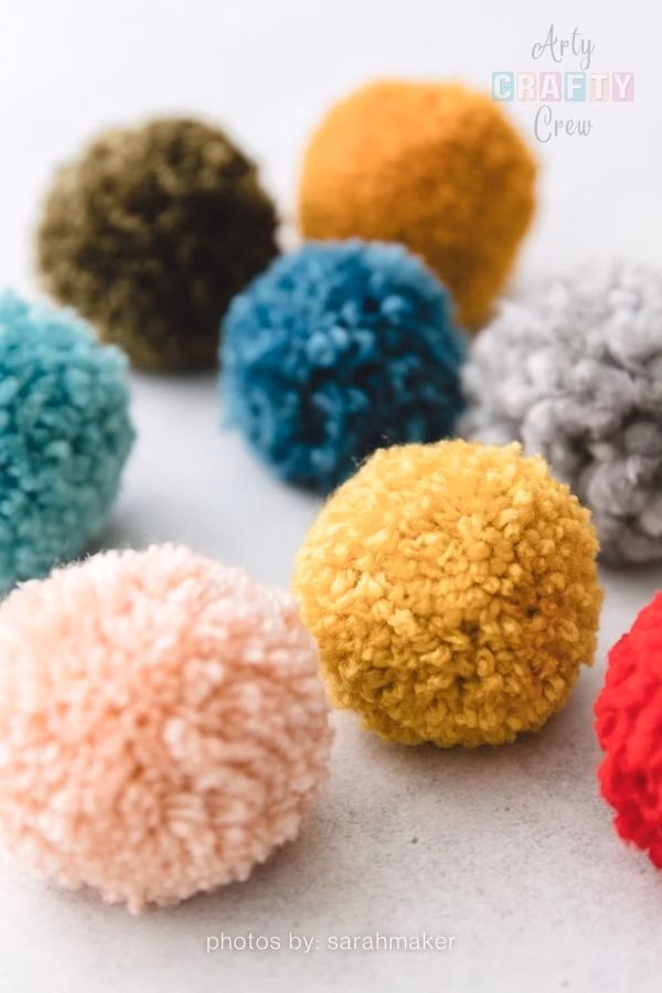 A close-up photograph showcasing a group of handmade, fluffy yarn pompoms resting on a smooth, light-colored surface. The pompoms are in a variety of solid colors, including pale pink, mustard yellow, dark teal, olive green, grey, and vibrant red. The Arty Crafty Crew logo is visible in the top right corner, and text at the bottom reads "photos by: sarahmaker". This is part of a craft roundup from https://artycraftycrew.com/.