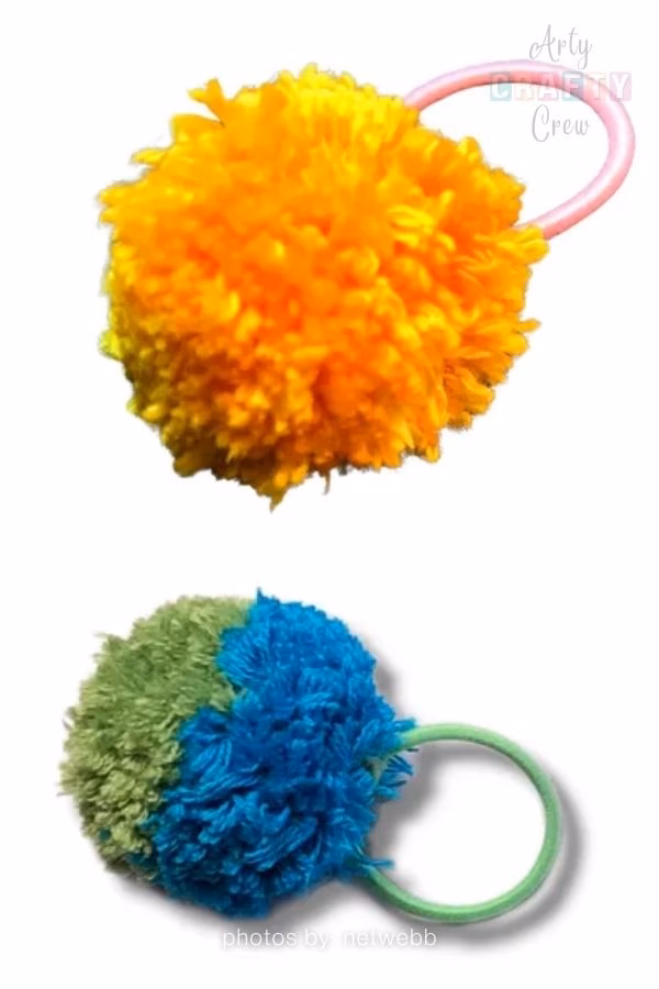 A vertically stacked image showing two handmade pompom hair ties against a plain white background. The top item features a large, fluffy orange and yellow pompom attached to a light pink elastic hair band. The bottom item features a two-toned fluffy pompom, split between olive green and bright blue, attached to a light green elastic hair band. The Arty Crafty Crew logo is visible in the top right corner, and text at the bottom reads "photos by: netwebb". This is part of a crochet roundup from https://artycraftycrew.com/.