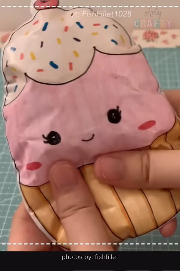 A close-up of a pink cupcake paper squishy with white frosting, colorful sprinkles, and a small cherry on top. The squishy features a smiling kawaii face and is shown being squeezed, featured in a tutorial collection from https://artycraftycrew.com/.