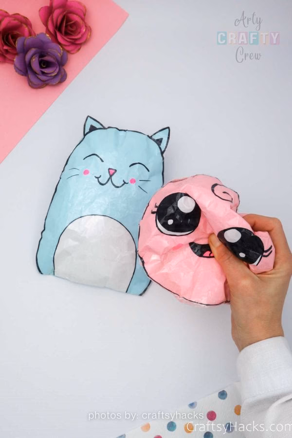 A top-down view of two handmade paper squishies: a light blue cat with a white belly and pink cheeks, and a pink flamingo being squeezed by a hand. These budget-friendly crafts are part of a DIY roundup from https://artycraftycrew.com/.