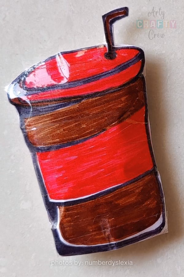 A handmade paper squishy designed to look like a takeaway coffee cup with a red sleeve and a straw. The craft has a glossy, laminated texture and is part of a budget-friendly DIY roundup from https://artycraftycrew.com/.