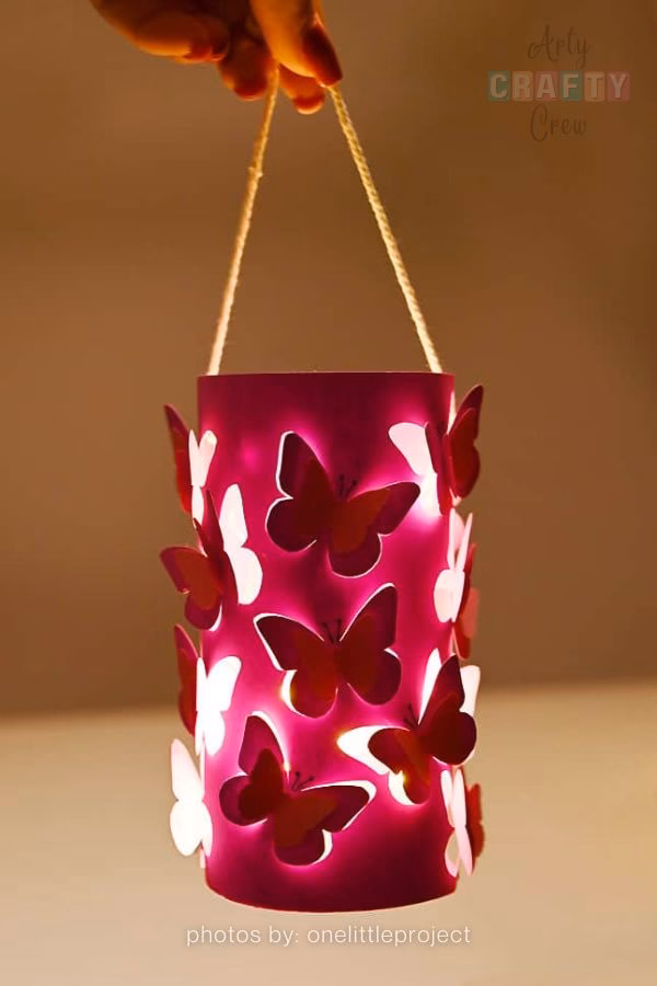A handmade pink cylindrical lantern with butterfly cut-outs that glow from an internal light source, featured in a craft roundup from https://artycraftycrew.com/. The lantern is being held by a string handle against a warm, dim background, highlighting the magical light effect through the butterflies.