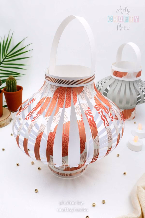 A pair of DIY paper lanterns featuring a modern orange and white patterned design, showcased in a craft roundup from https://artycraftycrew.com/. The lanterns have a classic rounded shape with a white handle and are displayed on a white surface alongside small potted plants and decorative gold beads.