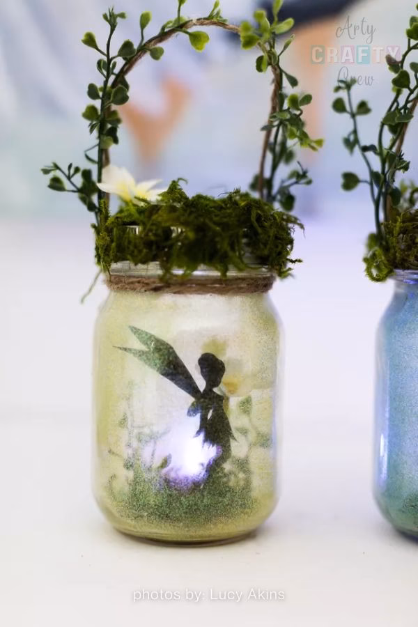 A whimsical DIY fairy lantern made from a mason jar, featured in a craft roundup from https://artycraftycrew.com/. The frosted jar contains a fairy silhouette and a soft light, topped with green moss and a decorative twine handle entwined with artificial greenery.